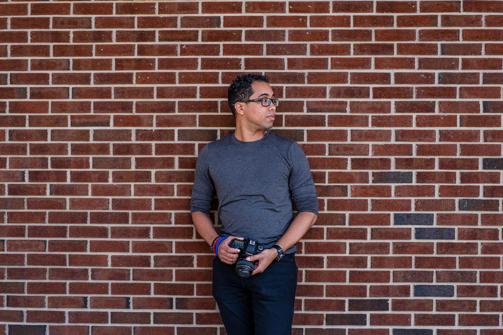 Casual male photographer standing with camera against brick wall, looking aside.